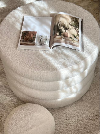 Polsterhocker als Ablagefläche – praktisches Designmöbel im Color-Blocking-Look Runder Polsterhocker in cremefarbenem Stoff mit aufgeklapptem Interior-Magazin im Color-Blocking-Stil