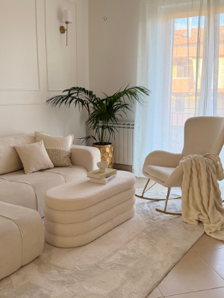 Beiges Wohnzimmer mit Sofa, passendem Hocker, gem&uuml;tlichem Sessel und Pflanze am Fenster