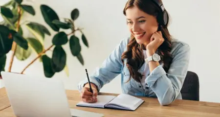 Junge Frau mit Headset am Laptop, l&auml;chelt und schreibt Notizen &ndash; symbolisiert digitales Arbeiten oder Online-Meeting