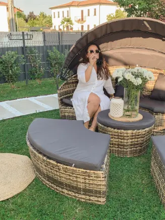 Person sitzt in einer runden Polyrattan-Gartenlounge mit Sonnenverdeck, grauen Auflagen und Beistelltisch im Garten